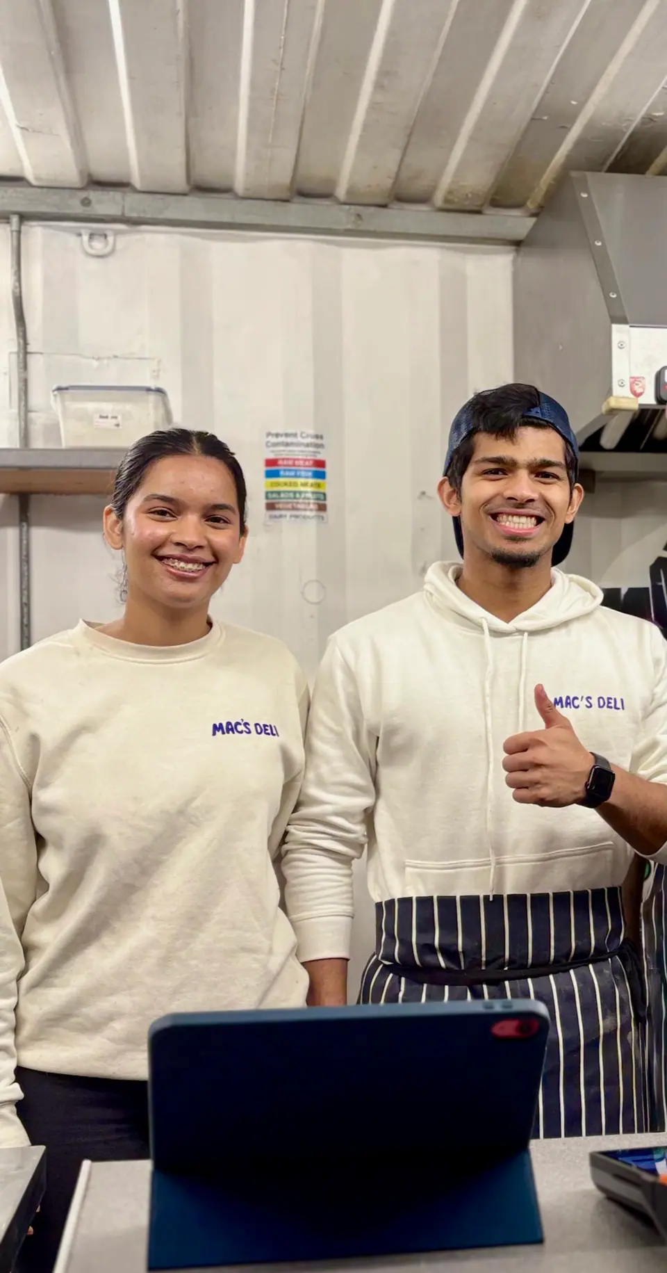 Two of the Mac’s Deli team in Mac’s Deli sweatshirts, smiling behind the counter