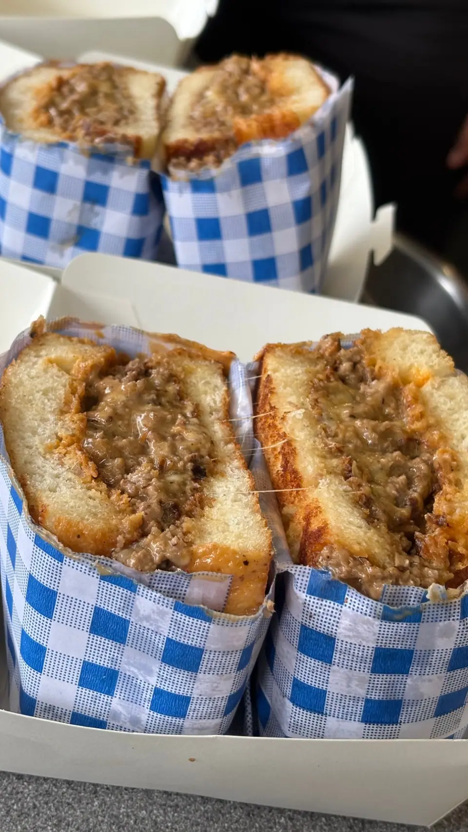 Two Mac’s Deli toasted sandwiches cut in half in blue gingham wrappers, packed in takeaway boxes