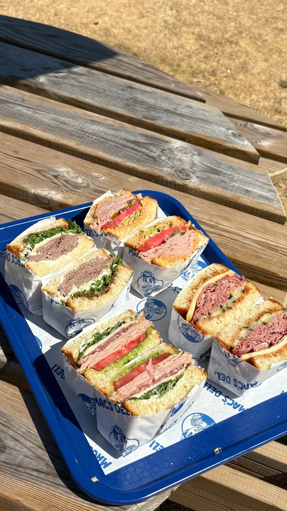 A blue tray of Mac’s Deli sandwiches cut in half in branded wrappers, on a sunny wooden picnic table